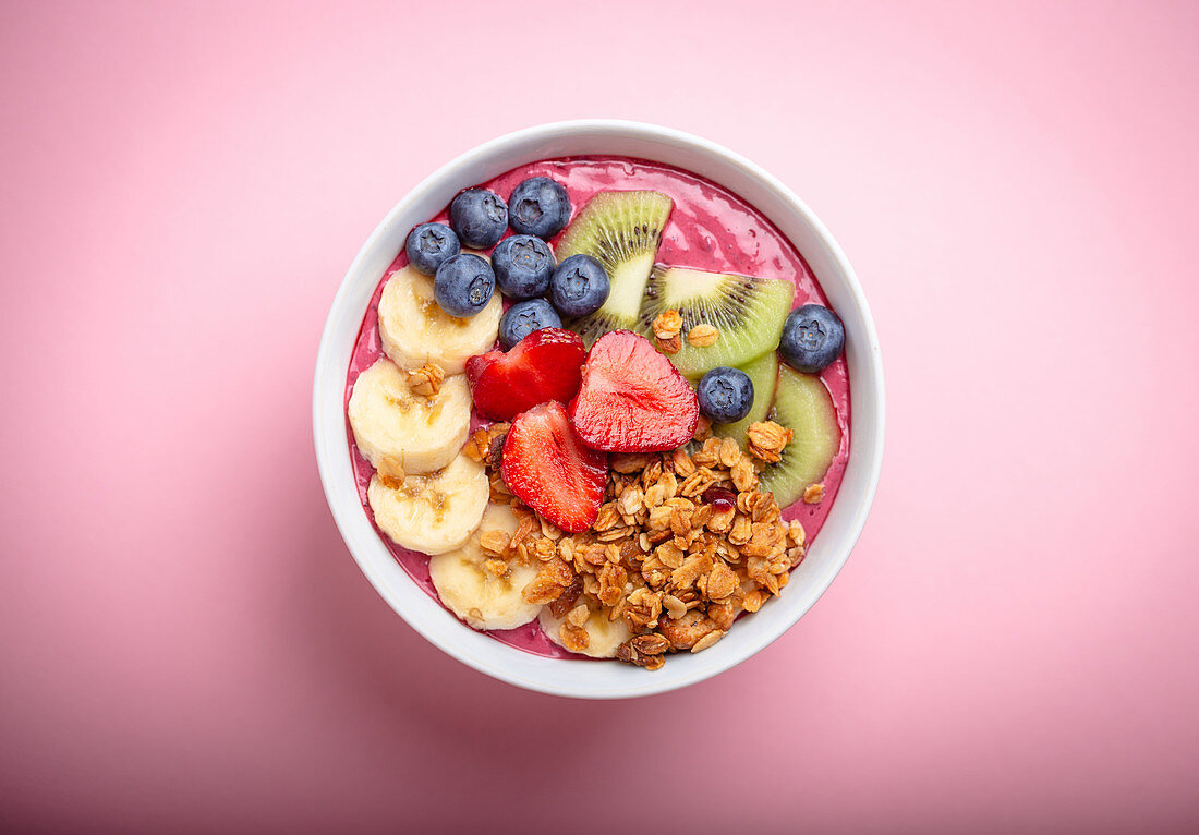 Summer acai smoothie bowl with strawberries, banana, blueberries, kiwi fruit and granola on pastel pink background
