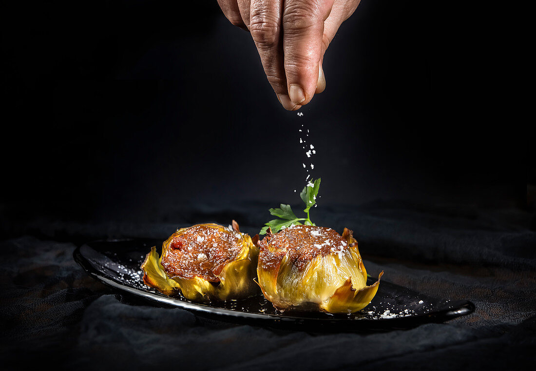 Person sprinkling tasty stuffed artichokes with salt