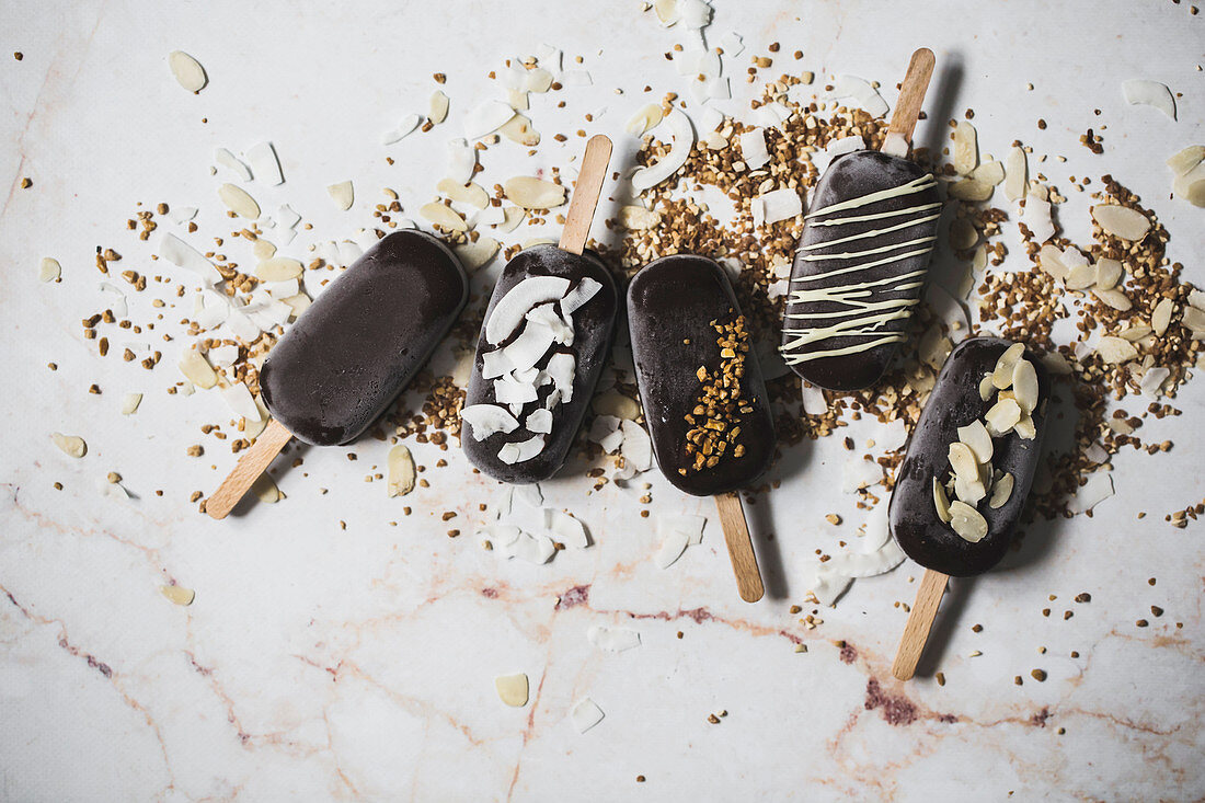 Assorted chocolate ice cream popsicles covered with different toppings on a marble surface