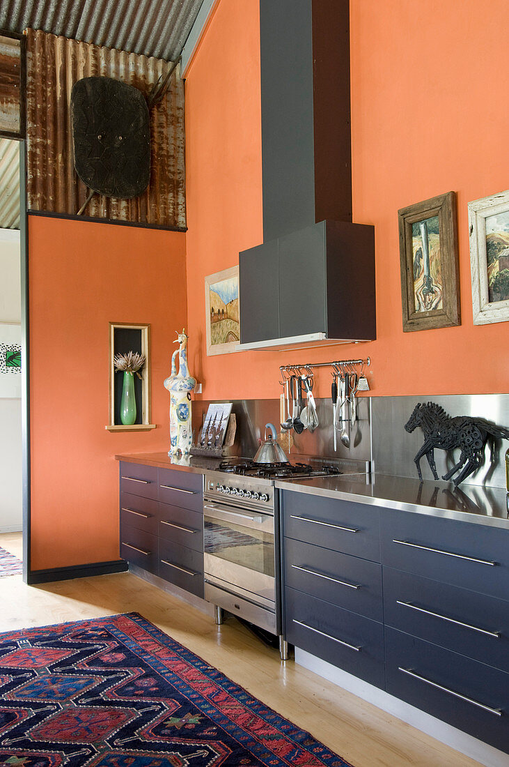 Modern kitchen counter with blue drawer fronts, stainless steel splashback and extractor hood in open-plan kitchen with orange walls