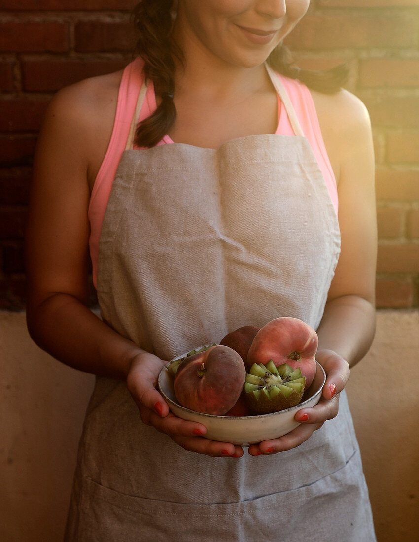 Tasty appetizing peaches and cut ripe juicy kiwi in bowl in hands of smiling woman in apron