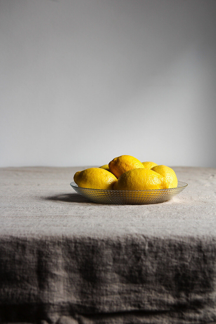 Appetizing lemons with shiny yellow peel in glass plate on grey linen tablecloth