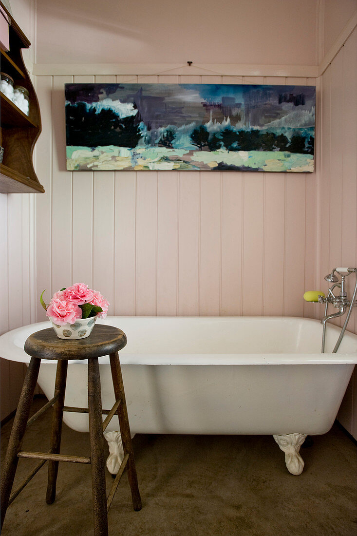 Vintage bathtub below painting in wood-clad bathroom with bowl of flowers on stool