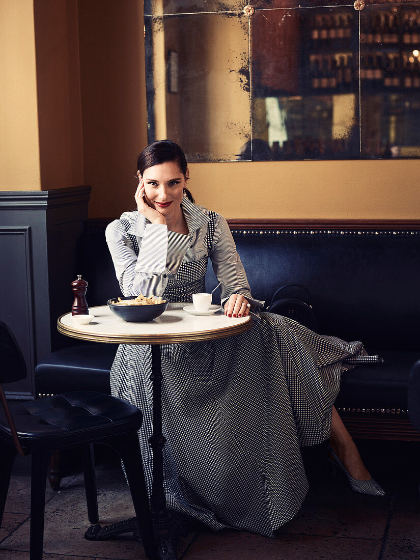 A dark-haired woman sitting in a restaurant wearing a striped shirt-blouse with a checked pinefore