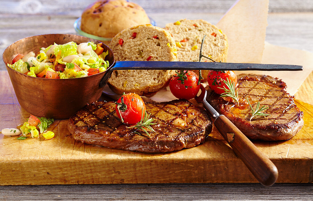 Grilled beef steaks with a vegetable salad and bread, cherry tomatoes, rosemary and a vintage copper pot
