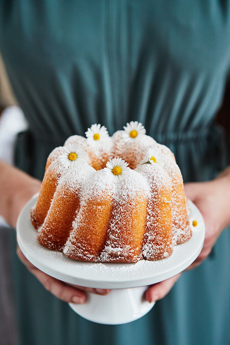 A woman holding a lemon Bundt cake decorated with daisies for a birthday