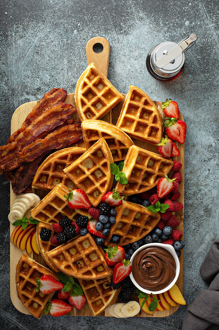 Big breakfast waffle bar served on a wooden board with berries, bacon and chocolate sauce