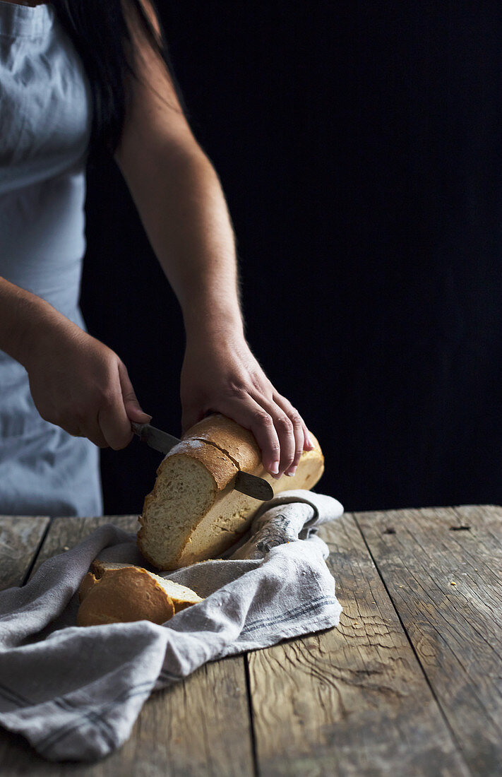 Unrecognizable female cutting loaf of fresh bread on napkin while standing near old wooden table on black background