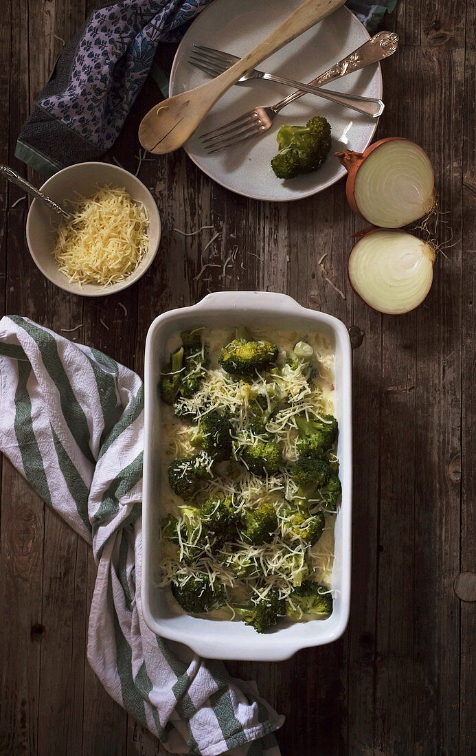 Ceramic baking pan of tasty roasted broccoli with shredded cheese and onion placed near napkin and dishware on timber tabletop