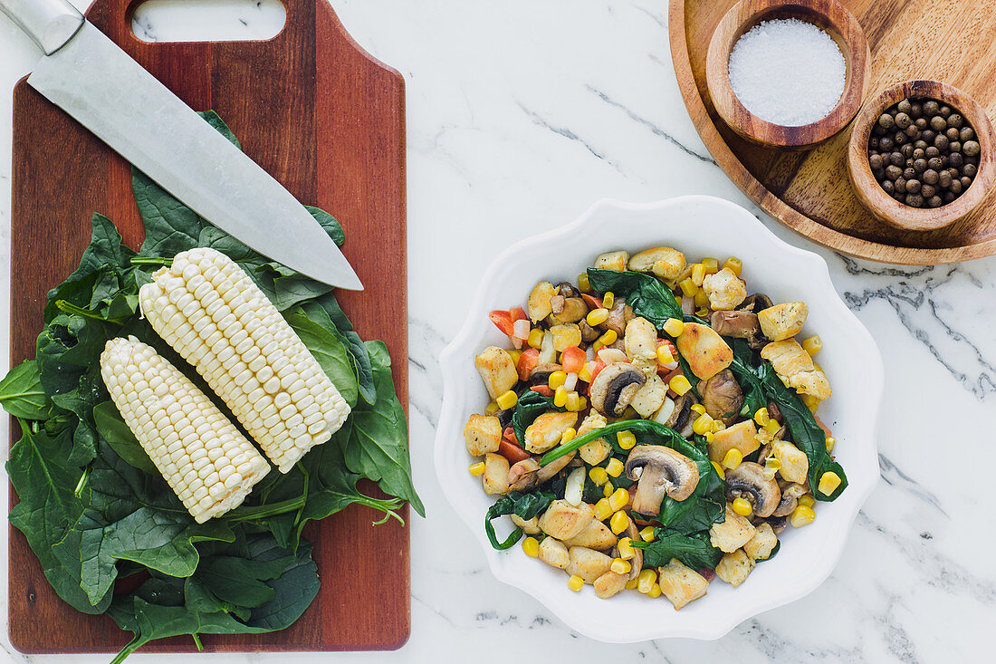 Served bowl with mushrooms corn and cut chicken on table with corn leaves of greenery on cutting board and condiments