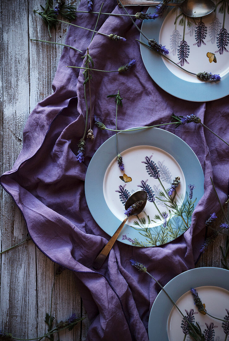 From above fresh lavenders placed around empty plates over violet fabric on lumber tabletop