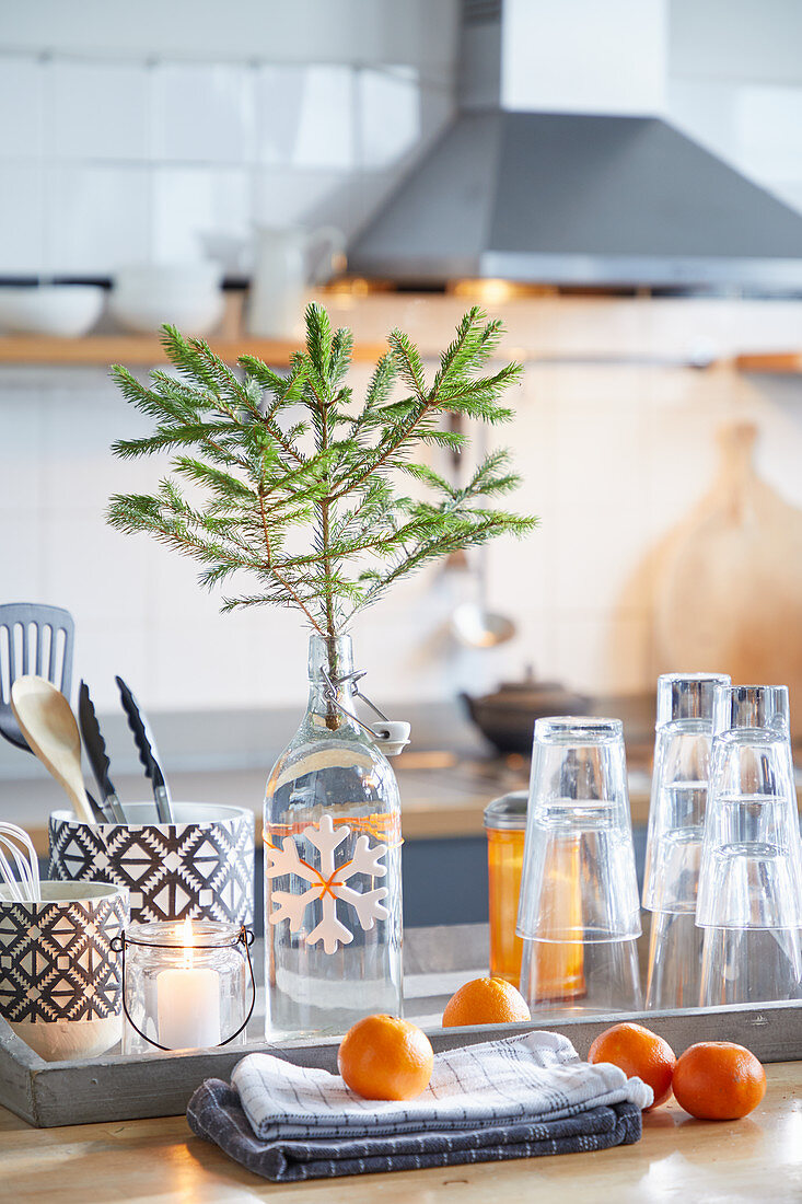 Fir branch in swing-top bottle. glasses and cutlery on tray behind tangerines on folded tea towels