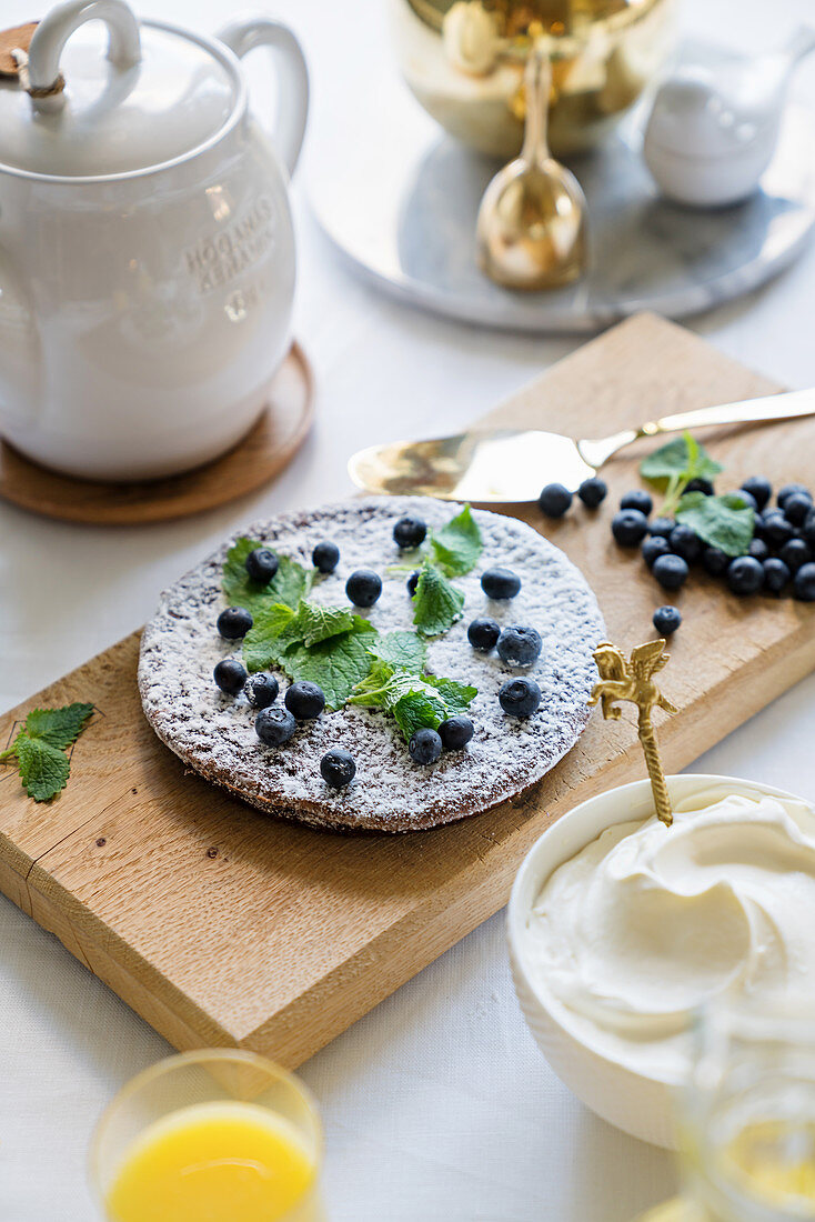 Bowl of whipped cream next to flat cake decorated with blueberries and lemon balm
