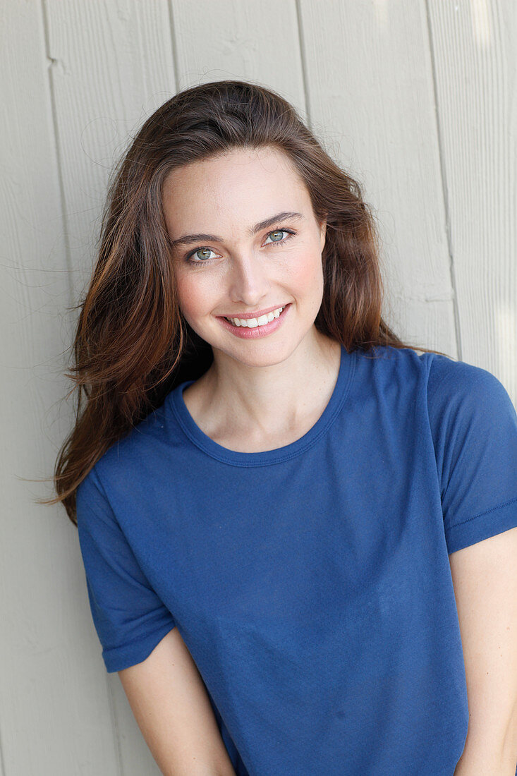 A young brunette woman in front of a grey wooden wall wearing a blue t-shirt