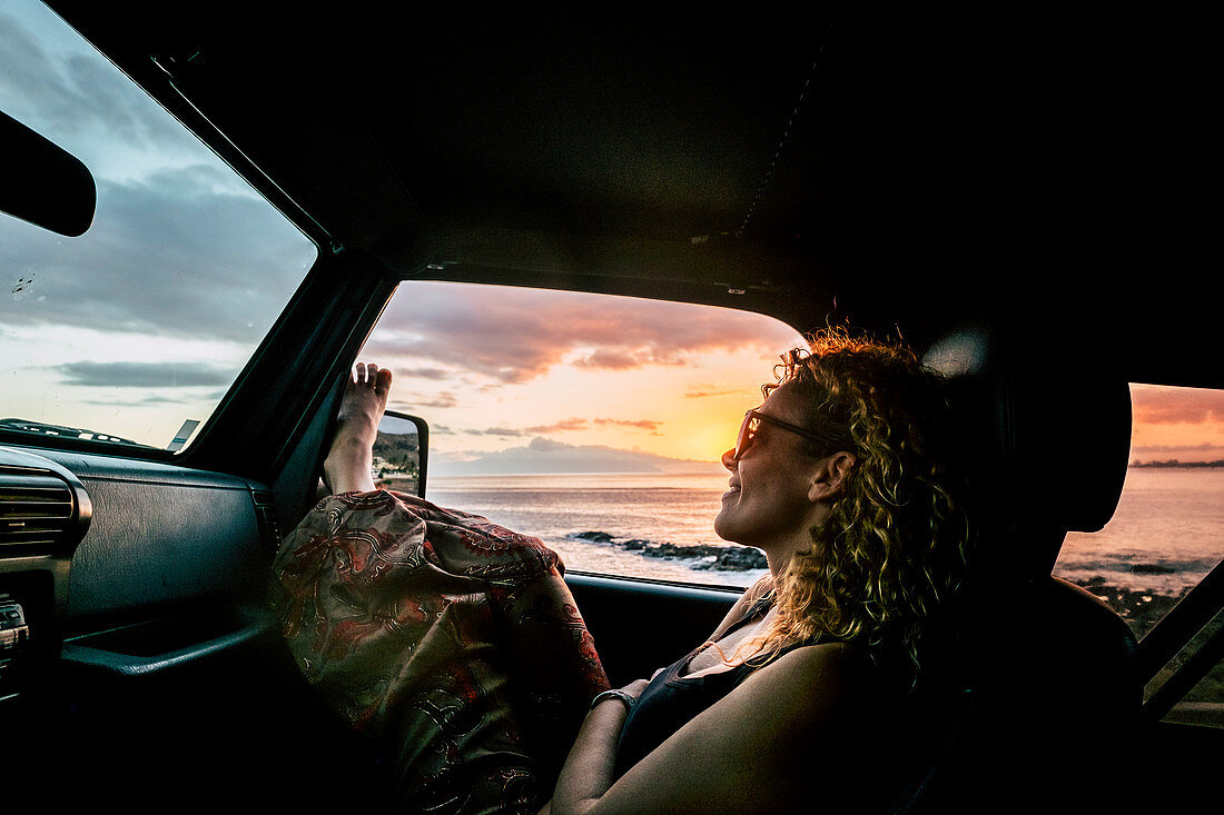 A woman sitting in a car at sunset with her feet out the window