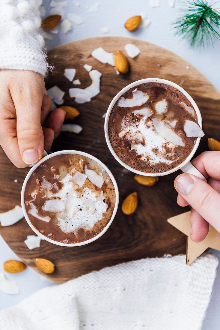 Hände halten zwei Tassen vegane heiße Schokolade mit Mandelmilch und Kokoschips