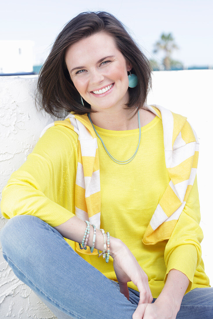 A young woman wearing a long-sleeved yellow top and jeans with a jumper over her shoulders