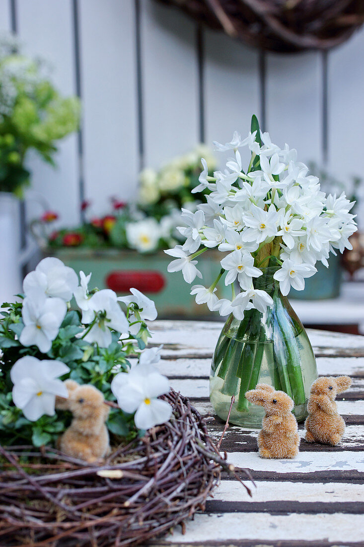 Little Easter bunnies, vase of narcissus 'Ziva' and white violas in wreath of twigs