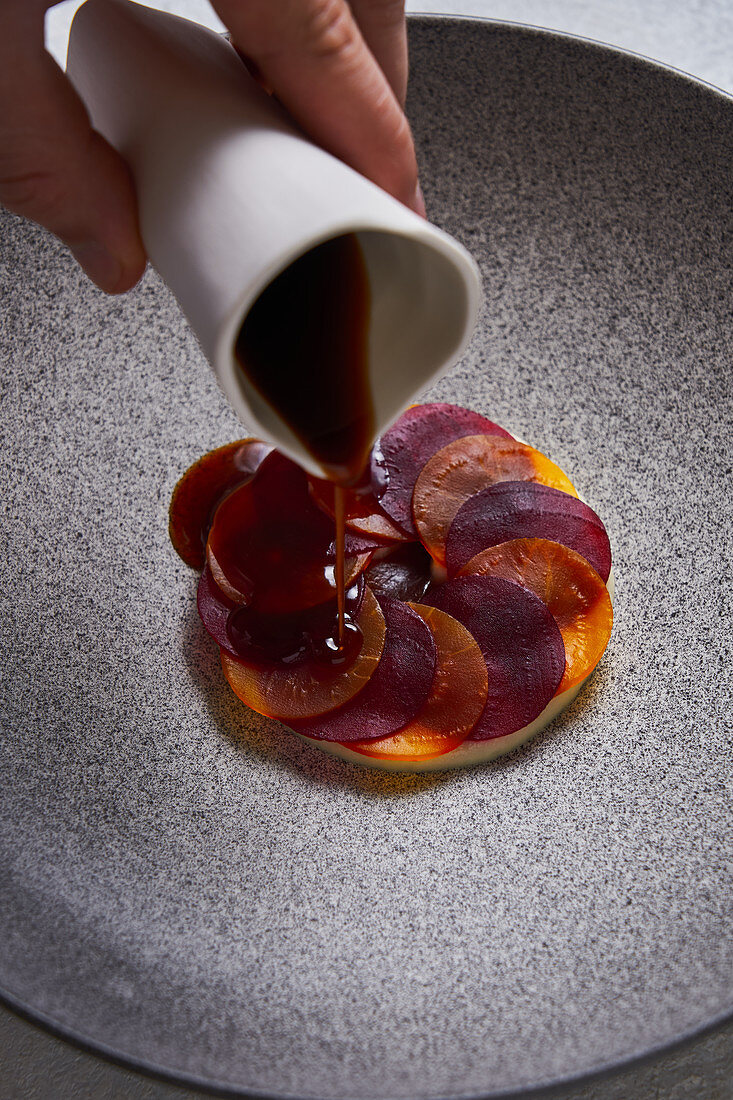 Person adding sauce to dish made of thin slices of boiled carrot and beetroot on plate