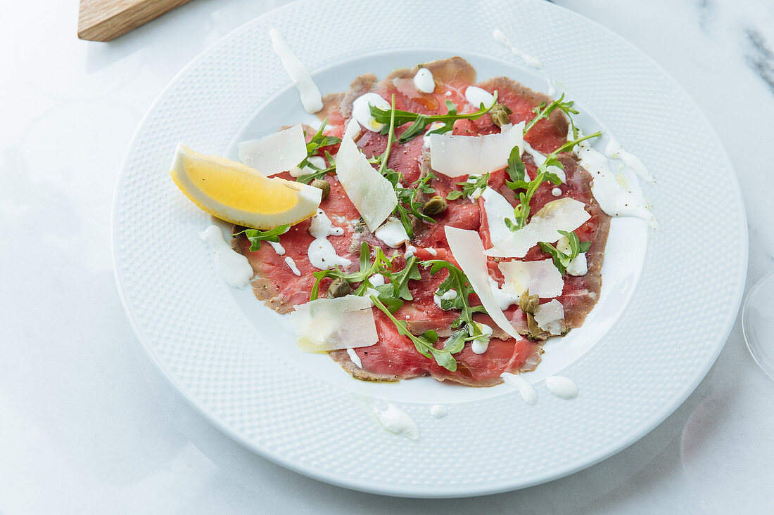 Slices red beef on plate with pieces of fresh lemon with white sauce cheese and arugula
