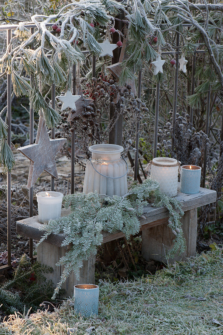 Christmas decoration on the garden fence with pine, rose hips and wooden stars, bench with frosted lanterns and a pine branch