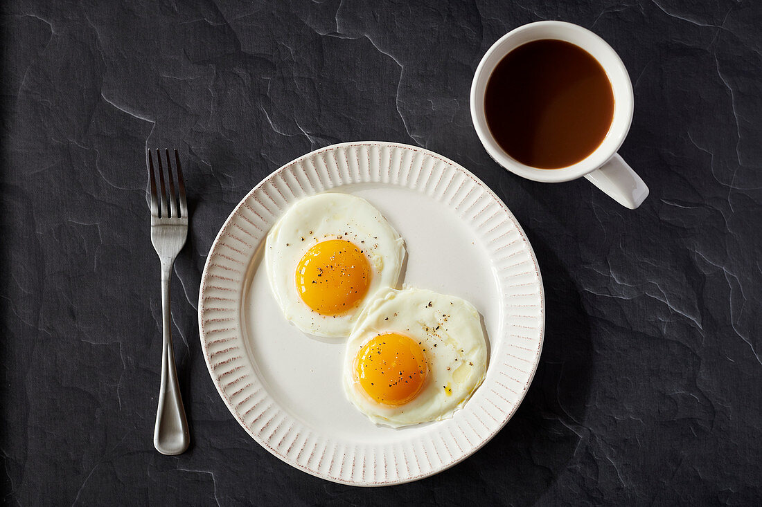 Two sunnyside eggs with pepper in a white plate with coffee in a white coffee cup on a black surface