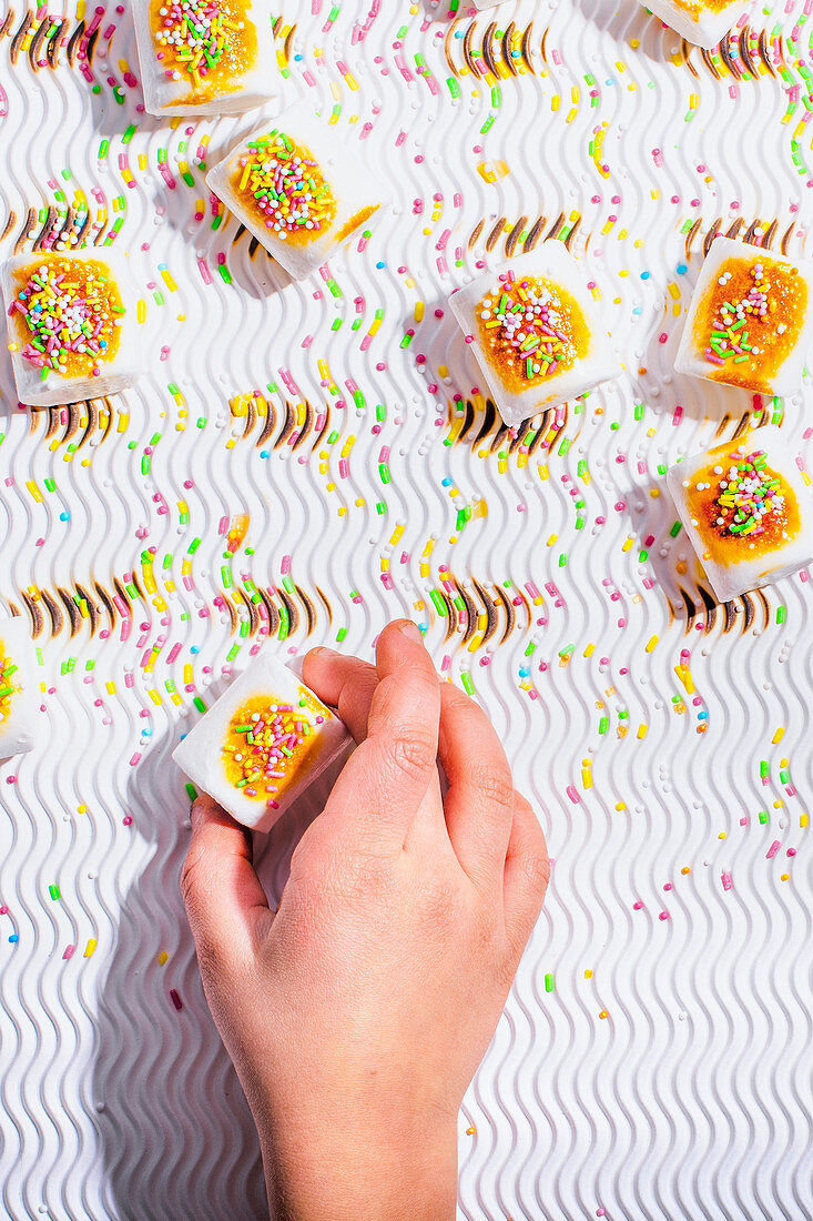 A child's hand reaches for a marshmallow with colorful sugar sprinkles