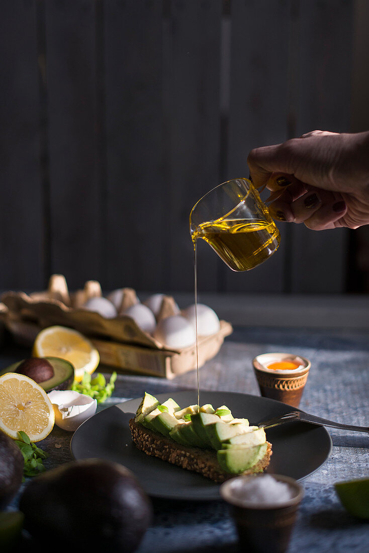 Olivenöl wird aus Glaskännchen auf Avocado-Brot geträufelt