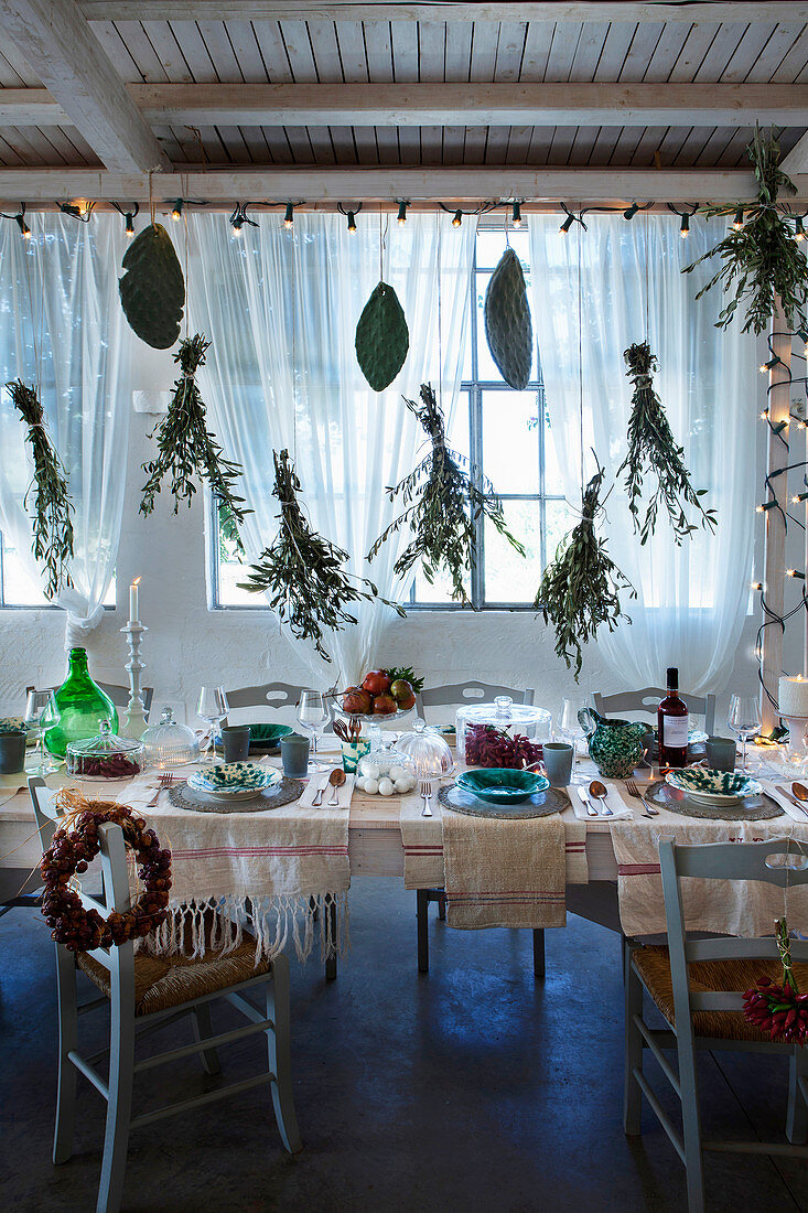 Festively decorated dining table below fairy lights and suspended bunches of leafy branches