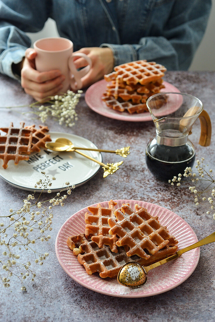 Waffles on a saucer on the table a jug of coffee a mug The woman is holding a cup in her hands