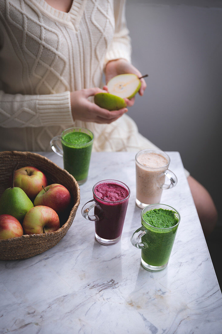Woman sitting at the table holding a pear in her hands and there are colourful smoothies in glasses on the table in front of her