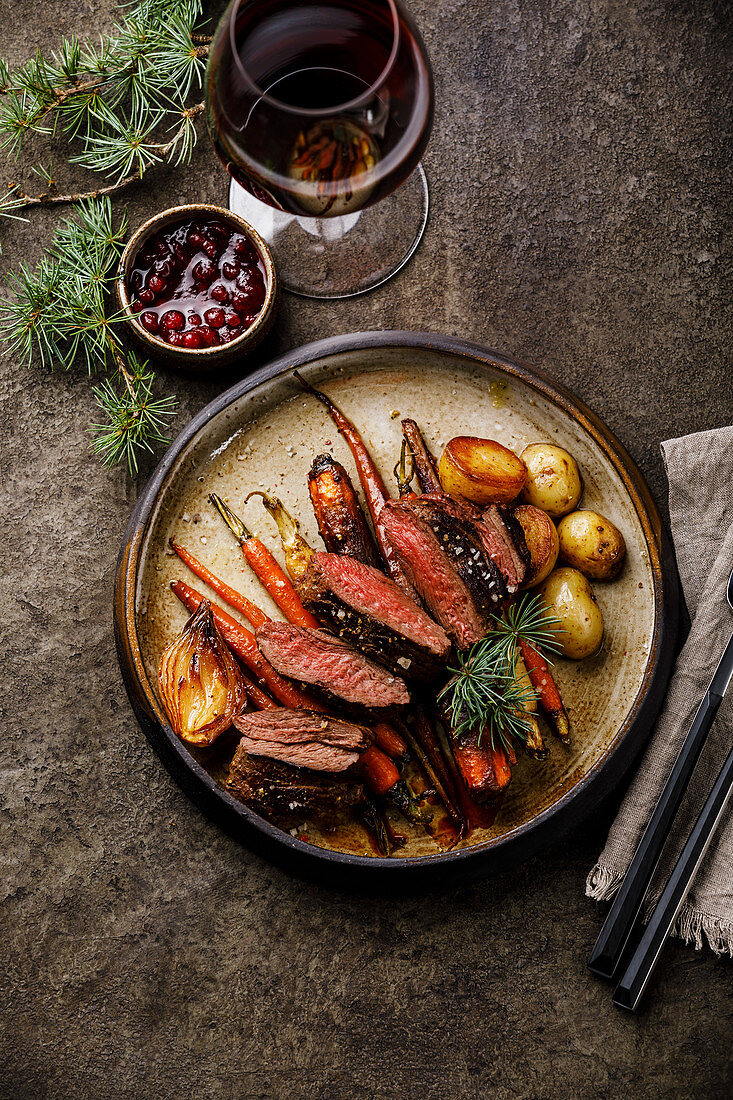 Grilled sliced Venison Steak with baked vegetables and berry sauce and Red wine on dark background