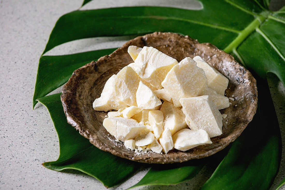 Chopped pieces of raw organic cocoa butter in craft ceramic plate on green exotic monstera leaf over grey spotted background. Close up