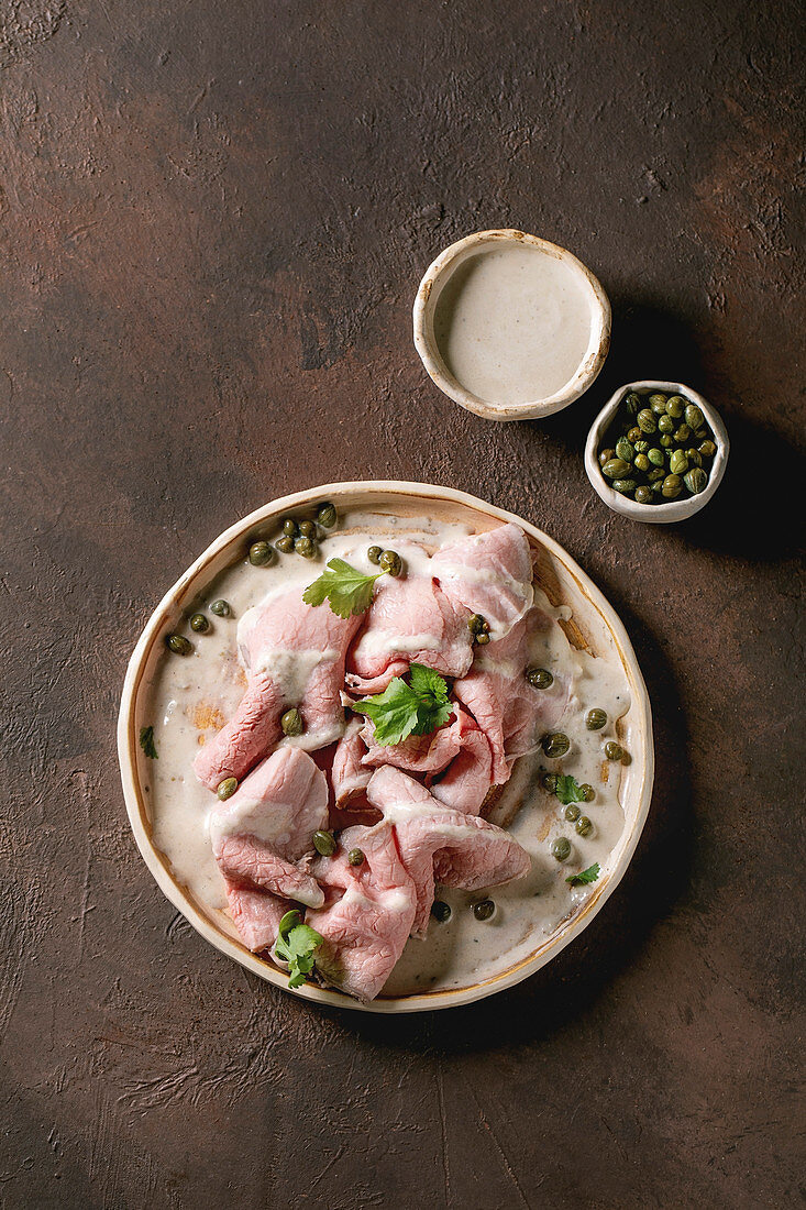 Vitello tonnato italian dish. Thin sliced veal with tuna sauce, capers and coriander served on ceramic plate over dark texture background. Top view, copy space