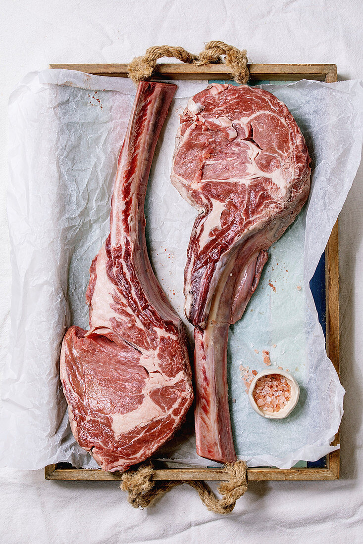 Raw uncooked black angus beef tomahawk steaks on bones served with pink salt on wooden tray with baking paper over white cloth as background. Top view, space.