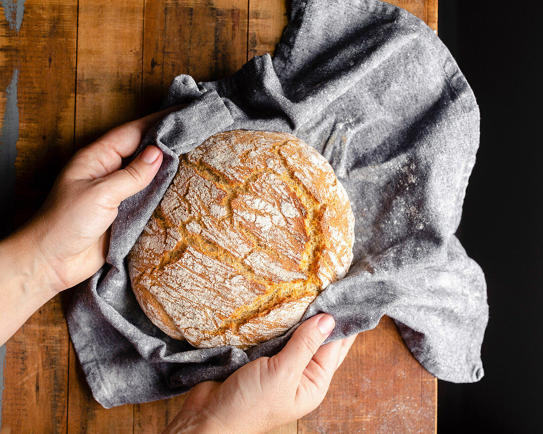 Hands placing a freshly baked loaf of bread wrapped in a grey cloth onto a wood tabletop.