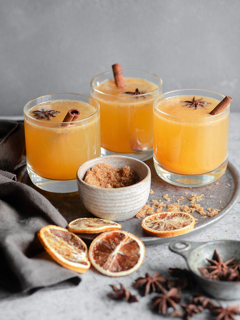 Three glasses of an orange spiced drink, garnished with cinnamon sticks, brown sugar, blood oranges, and star anise