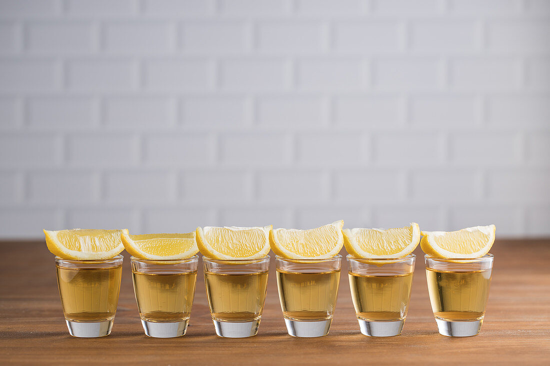 Glass sots with golden tequila and slices of lemon on wooden table