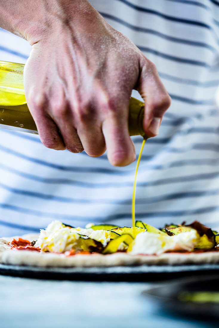 Olivenöl wird auf Pizza mit Zucchini, Mozzarella und Tomatensauce geträufelt