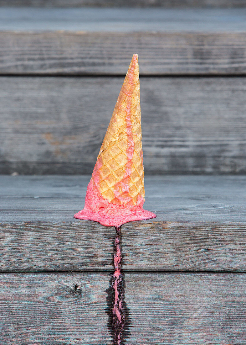 Pink ice cream in wafer cone dropped on wooden plank table with melting traces