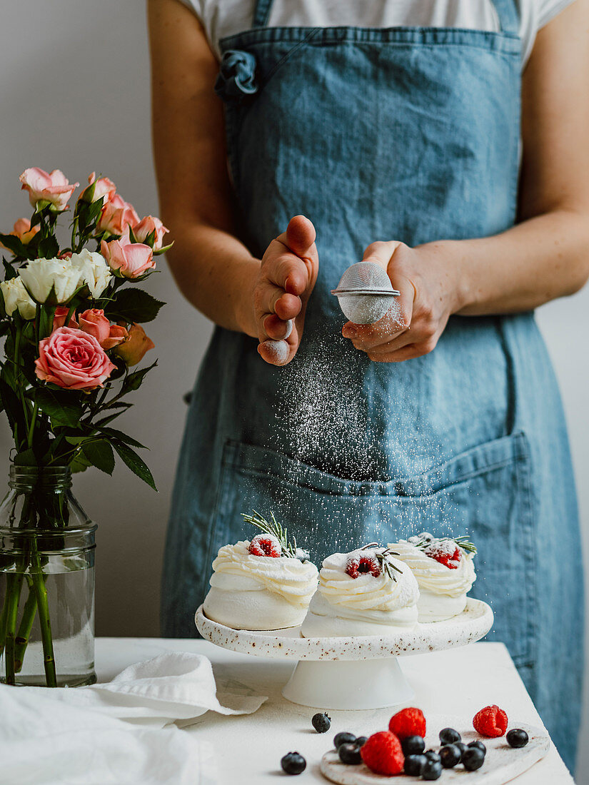 Woman sprinkles icing sugar on mini Pavlova cakes with fresh berries