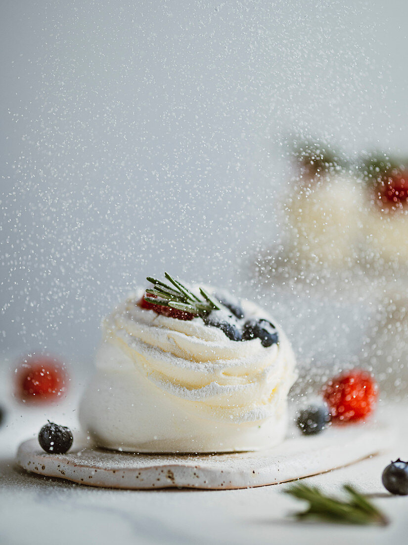 Icing sugar sprinkles on mini Pavlova cake with fresh berries and fresh green rosemary