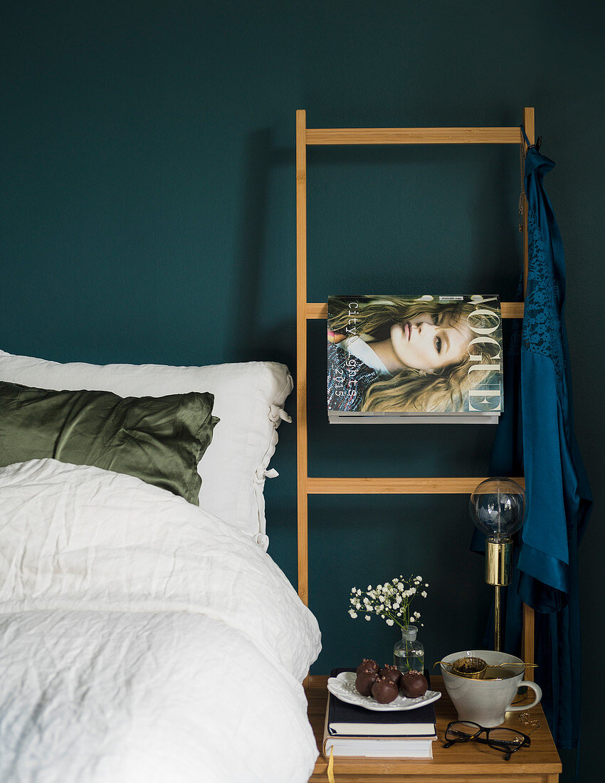 Bed with white bed linen and ladder used as magazine rack against petrol-blue wall