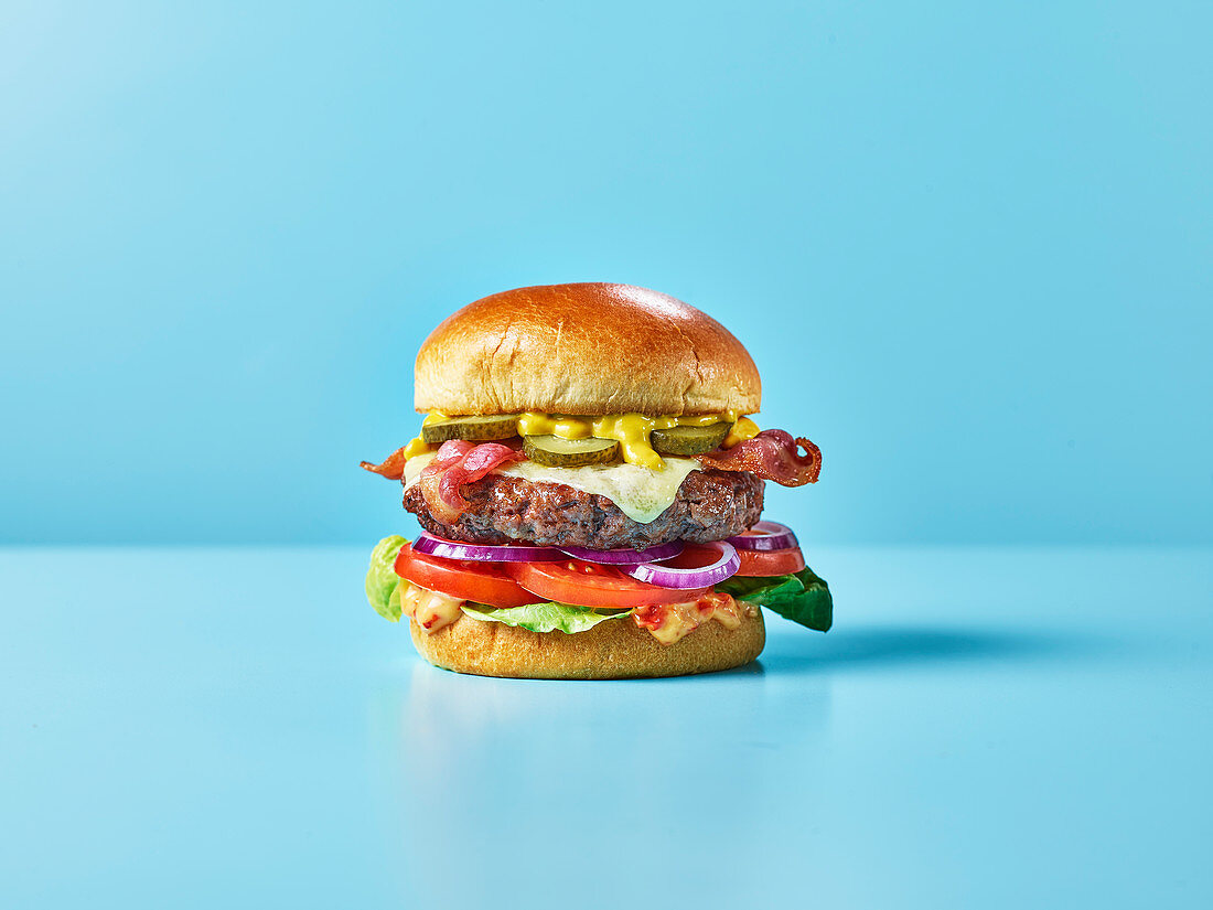 Cheeseburger mit Speck, Salat, Relish und Essiggurken vor blauem Hintergrund