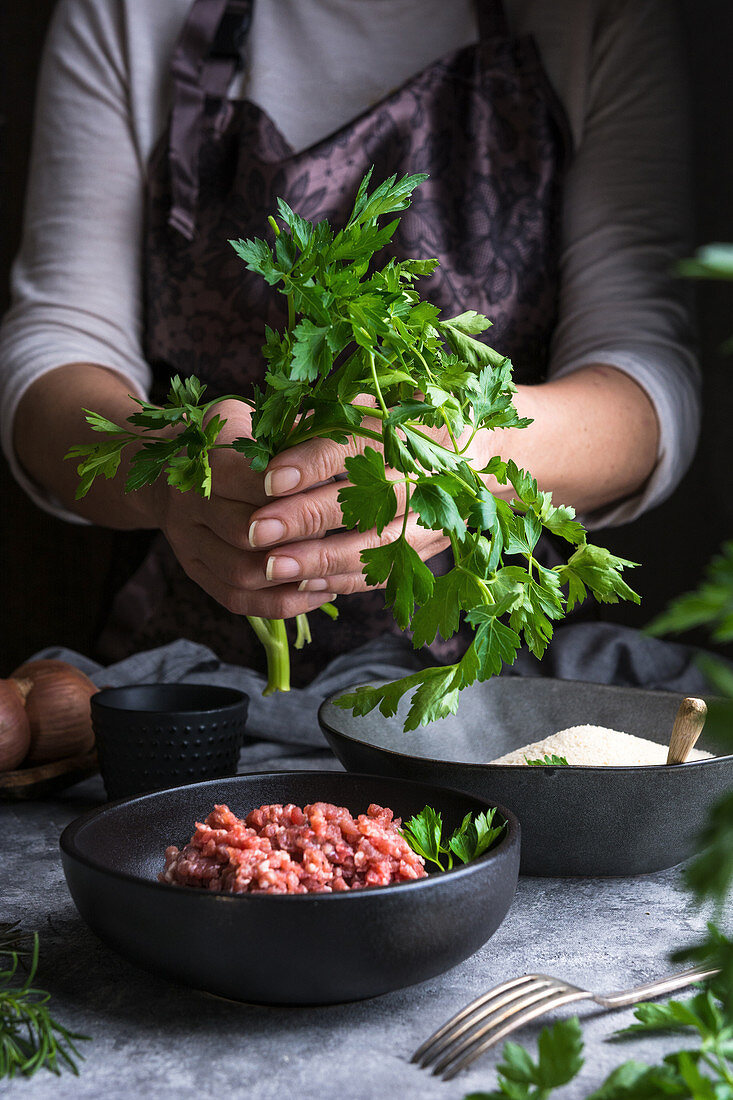 Unrecognizable female wearing apron keeping bunch of fresh parsley over bowl with raw minced meat