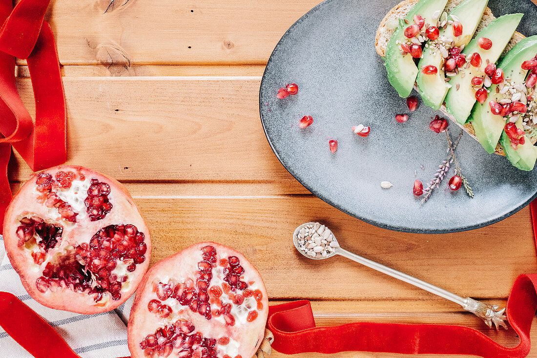 Halved pomegranate and spoon with seeds placed near plate with delicious avocado toast on wooden table