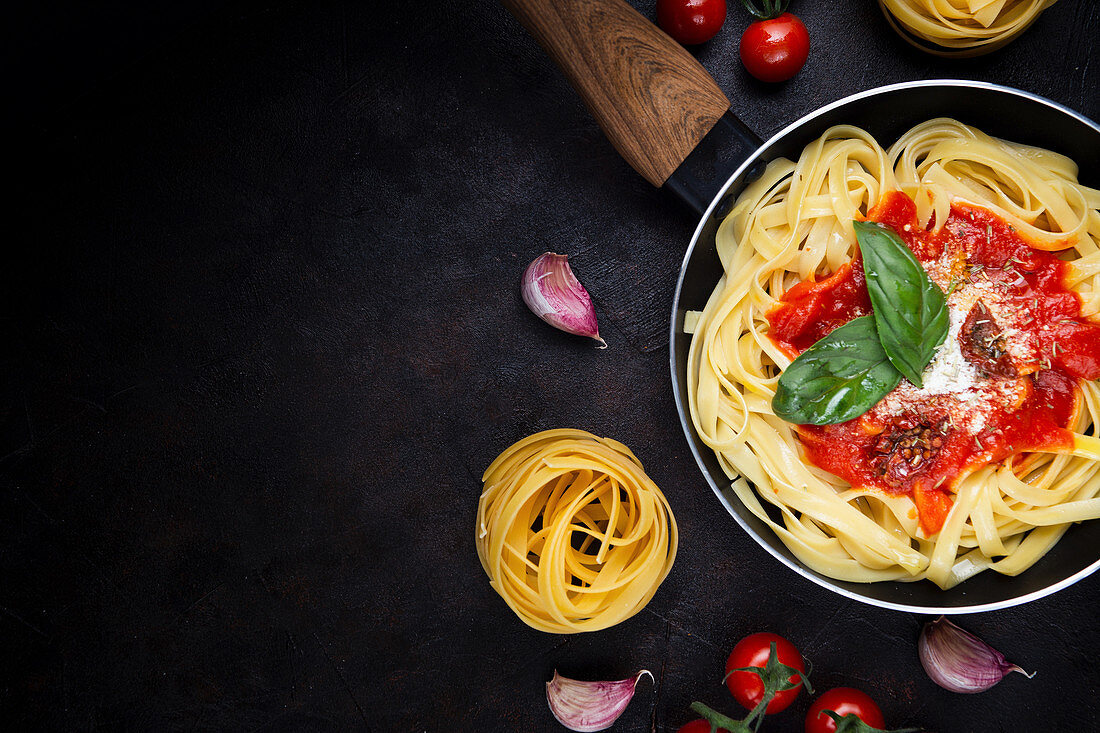 Tagliatelle pasta served in pan with tomato sauce and fresh basil leaves on black background