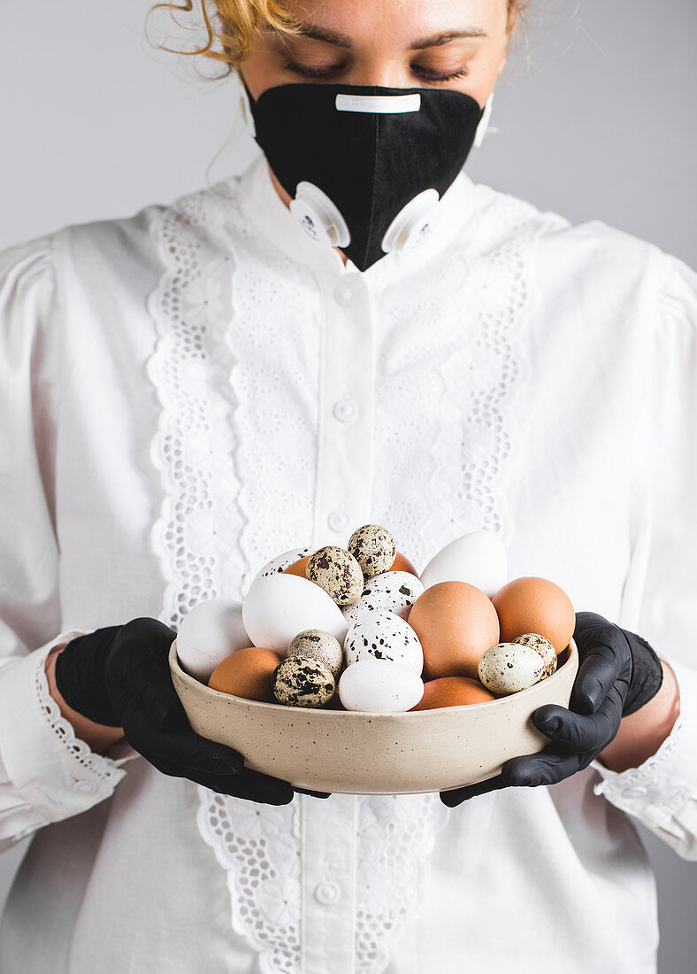 Frau in weißer Bluse mit schwarzer Gesichtsmaske und schwarzen Handschueh hält Schüssel mit Eiern