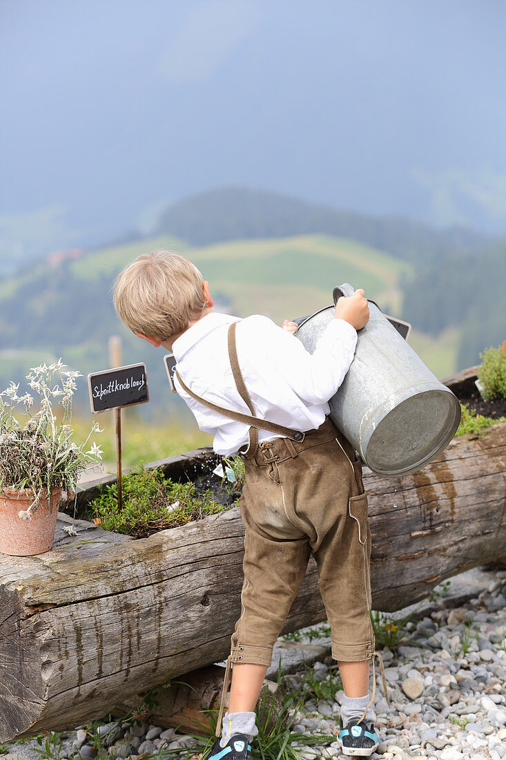 A little boy wearing lederhosen watering herbs on a mountain
