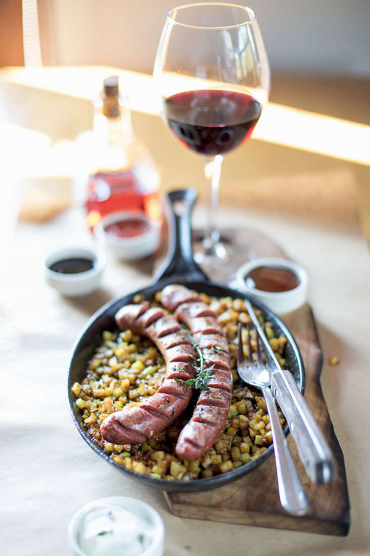 Sausages with Potatoes in a cast iron pan, Table served with glass of red wine
